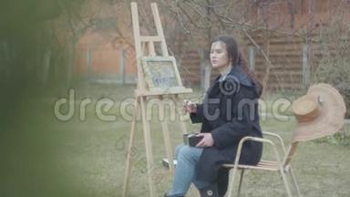 相当集中的年轻女画家坐在木制画架前画一幅画。 休闲女艺术家